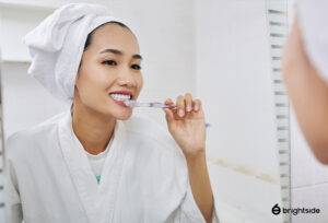 Woman smiling and brushing teeth in the mirror with towel on head