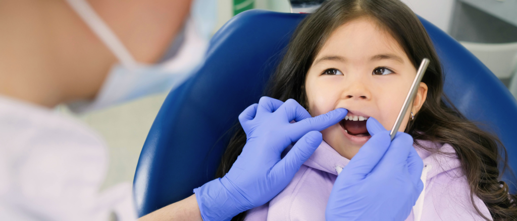 A child sitting in a dentist's chair, looking curious and calm, while the dentist, wearing gloves and a mask, gently examines their teeth. The brightly lit dental clinic is clean and inviting, with tools neatly arranged on a tray nearby.
